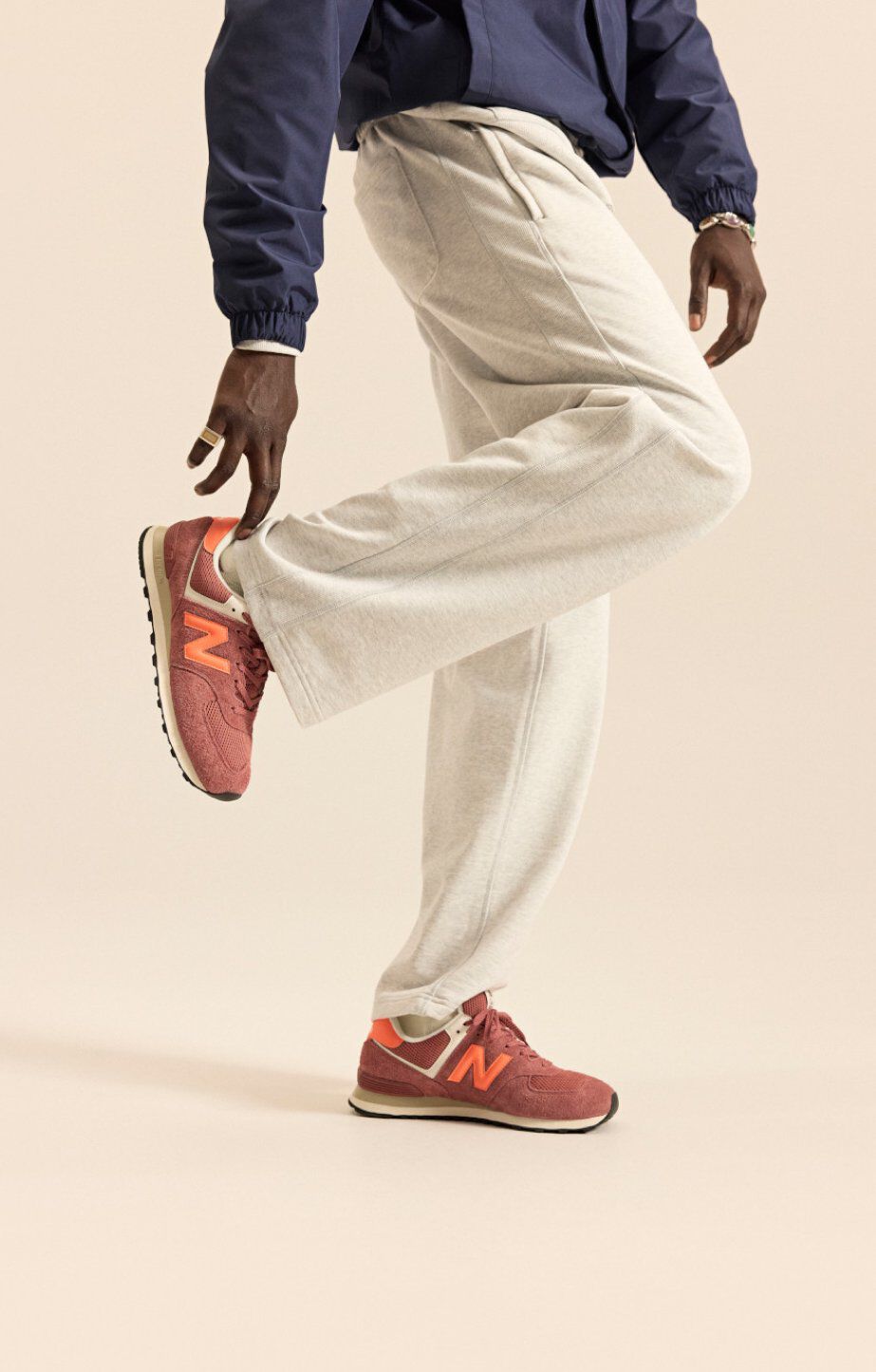 Person wearing brick‑red New Balance sneakers with orange accents, shown mid‑pose against a neutral background. 